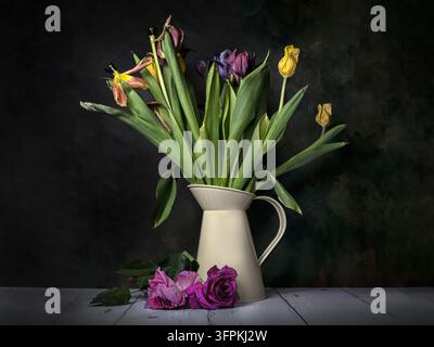 Stillleben mit verblassten Tulpen in einer Kanne mit toten Rosen auf weißem Holztisch vor dunklem Hintergrund Stockfoto