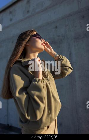Stylische junge Frau trägt einen olivgrünen Hoodie und passt trendige Sonnenbrille im Freien an einer Betonwand bei hellem Sonnenlicht an. Moderne urbane Mode Stockfoto
