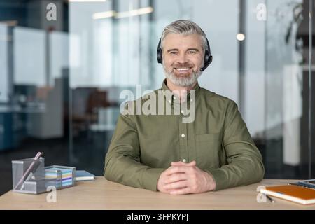 Selbstbewusster männlicher Callcenter-Agent oder Kundendienstmitarbeiter, der lächelnd an einem Schreibtisch in einem zeitgenössischen Büroumfeld sitzt und professionelle Online-Unterstützung und Kommunikation anbietet Stockfoto