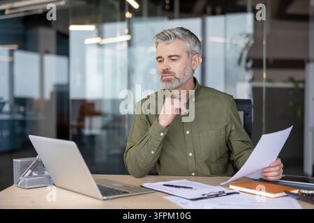 Erfahrener Geschäftsmann mit grauen Haaren und Bart sitzt an einem Bürotisch, überprüft Finanzberichte und arbeitet an einem Laptop, zeigt Konzentration und berufliche Verpflichtung in einem modernen Arbeitsumfeld Stockfoto
