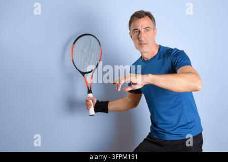 Ein Mann mittleren Alters hält einen Tennisschläger, während er sich zum Schlag bereitmacht, und streckt seinen freien Arm zur Balance in einem dynamischen Studio-Sportporträt aus. Stockfoto
