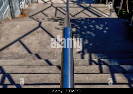 Ein vertikales Edelstahl-Geländer, das in der Mitte einer Reihe von Beton-Außenstufen verläuft und geometrische Schatten über die Stufen wirft. Stockfoto