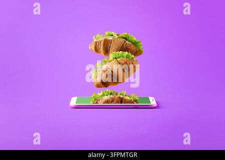 Konzept des Online-Lebensmittellieferdienstes mit Kopierraum. Fliegende Sandwiches von einem Smartphone auf einem lila Hintergrund. Stockfoto