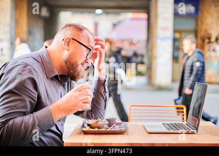 Mann hält ein Getränk und berührt den Kopf, während er einen Laptop in einem Café im Freien benutzt, zeigt Stress oder Müdigkeit während der Fernarbeit, Konzept von Überlastung und Work-Life-Balance im modernen Lebensstil Stockfoto