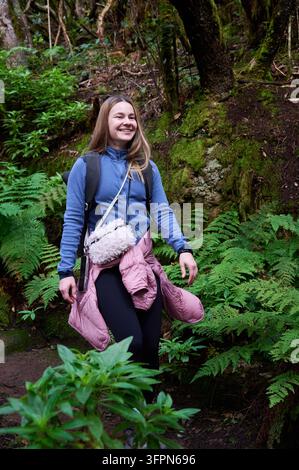 Junge Frau wandert durch dichten, von Farnen bedeckten Wald mit Rucksack, lächelt, während sie auf einem unebenen Waldweg unter moosigen Bäumen in den Anaga-Bergen, Teneriffa, Kanarische Inseln, Spanien geht Stockfoto