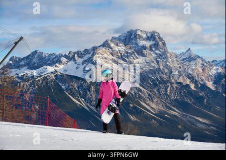 Snowboarderin steht auf schneebedecktem Hang in den Dolomiten, Italien, hält ihr Snowboard neben sich und posiert vor den schroffen Alpen Gipfeln Punta Sorapis während eines Wintersporttripps und eines aktiven Bergurlaubs Stockfoto