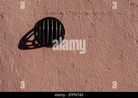 Ein schwarzer Metallwandlüfter mit vertikalen Stäben wirft einen langen Schatten auf eine raue, staubige rosa Stuckfläche im warmen Sonnenlicht. Stockfoto