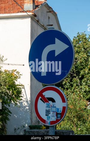 Blueschild für Rechtsabbiegen über einem mit Aufklebern bedeckten Schild für ‚Keine Linksabfahrt‘ neben einem alten Haus und grünen Bäumen an einem sonnigen Tag Stockfoto