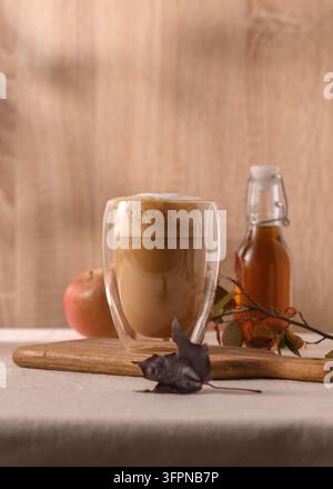 Schaumiger Apfel-Ahorn-Latte in einem klaren Glas auf einem Holzbrett, Apfel- und Sirupflasche im Hintergrund, Herbstblätter schaffen eine gemütliche Stimmung. Stockfoto