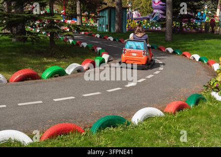 Frau und Kind fahren ein kleines orangefarbenes Elektroauto entlang einer kurvenreichen Strecke, die von roten, grünen und weißen Reifen gesäumt ist, in einem sonnigen Park. Stockfoto
