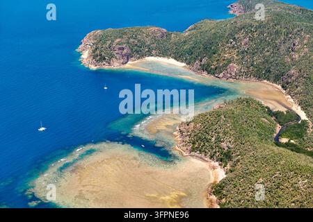 Segelboote, die in der Nähe einer tropischen Inselbucht vor Anker liegen, wo ein flaches Riffplateau und ein sandiger Strand auf bewaldete felsige Landspitzen in Australien treffen. Stockfoto