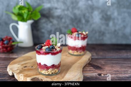 Schichtdessert mit Mascarpone, frischen Beeren und Granola, serviert in einem Glas und stilvoll auf einem rustikalen Holztisch angerichtet, mit zusätzlichem Beerendessert im Hintergrund Stockfoto