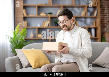 Junger Mann sitzt zu Hause auf einem Sofa, empfängt und öffnet eine kleine Pappschachtel mit einem glücklichen und überraschten Gesichtsausdruck, genießt den Moment des Auspackens einer neu angekommenen Online-Shopping-Bestellung Stockfoto