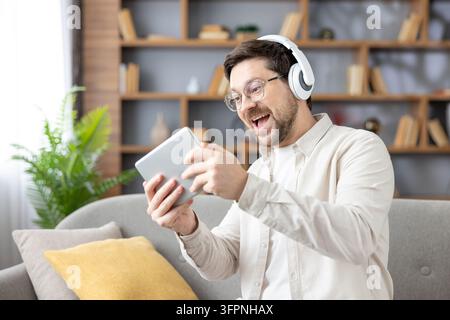 Mann mit Kopfhörern und Brille sitzt bequem auf einer Couch, vertieft ins Spielen eines Videospiels auf einem digitalen Tablet, feiert einen aufregenden Moment mit offenem Mund und freudiger Miene Stockfoto