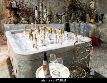Rustikale Weinkellerszene mit Champagnerflaschen, die in einer antiken Badewanne mit Eis gekühlt werden, neben einem verzierten Metallstuhl und einem Tisch, der für eine Verkostung gedeckt ist. Stockfoto