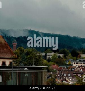 Deutschland. Bewölkte Ansicht einer ruhigen Hügelstadt mit Kirchturm, roten Dächern, Bäumen und Nebel, der über dunkle bewaldete Kämme zieht und ruhige Stimmung erzeugt Stockfoto