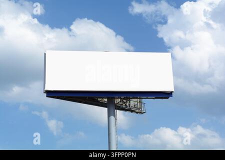 Leere weiße Werbetafel erhebt sich über offenem blauem Himmel mit sanften Wolken und lässt freien Kopierraum für Outdoor-Werbemockups. Stockfoto