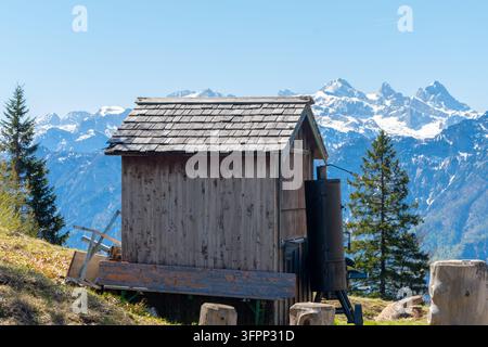 Eine kleine rustikale Holzhütte steht vor schneebedeckten Alpen und immergrünen Bäumen. Stockfoto