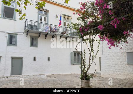 Rosa Bougainvillea umrahmt einen stein gepflasterten Innenhof und ein weiß getünchtes Gebäude mit grauen Fensterläden und einem eisernen Balkon in Hydra, Griechenland Stockfoto