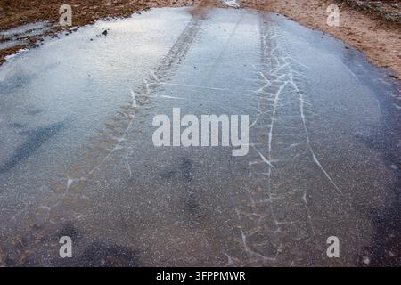 Autoreifenspuren, die sich in einer dünnen Schicht gefrorenen Pfützenwassers mit kunstvollen Rissmustern auf einer schlammigen Landstraßenoberfläche im frühen Winter abzeichnen Stockfoto