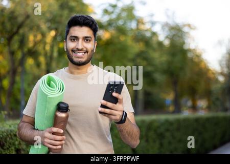 Junger Mann lächelt, während er eine gerollte Yogamatte, eine Wasserflasche und sein Smartphone hält, bereit für ein Outdoor-Workout und verbunden mit der Technologie in einer grünen Parkumgebung Stockfoto