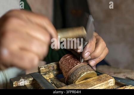 Eine Nahaufnahme der Hände eines Handwerkers, der mit traditionellen Handwerkzeugen akribisch komplexe Muster in einen kleinen Kupferbecher ein graviert Stockfoto