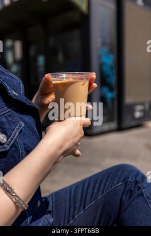 Nahaufnahme von weiblichen Händen, die einen Eiskaffee mit Milch halten, Frau in einer Jeansjacke mit einem funkelnden Armband, unscharfer Ladenfront Stockfoto