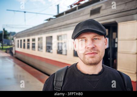 Männlicher Reisender mit Rucksack, der auf dem Bahnsteig neben einem Personenzug vor der Abfahrt steht. Stockfoto