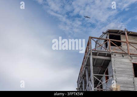 Niedriger Blick auf einen verwitterten grauen Holzturm mit rostigem orangefarbenem Geländer und einem Segelvogel, der in einem wolkigen blauen Himmel schwebt Stockfoto