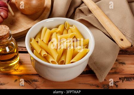 Ungekochte Penne-Pasta in einer weißen Keramikschüssel auf einem rustikalen Holztisch, begleitet von Olivenöl, Knoblauch und Zwiebel. Vorderansicht. Stockfoto