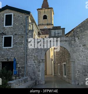 Ein steinerner Torbogen in Kroatien führt zu einer schmalen, sonnenbeschienenen Gasse zwischen verwitterten Mauerwerksgebäuden mit verschlossenen Fenstern und einem Kirchturm. Stockfoto