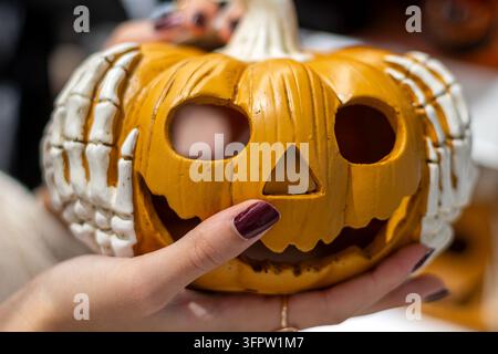 Nahaufnahme von weiblichen Händen, die eine orangefarbene Halloween-Kürbisknisterlaterne mit Skelettfingern halten, gruseliges Jack-O-Laternen-Gesicht mit geschnitzten Augen und einem Lächeln für Stockfoto