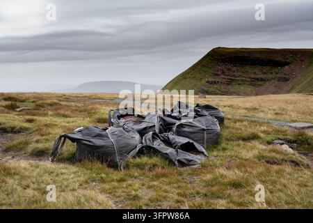 VERWORFENE BAUSÄCKE IN EINER BERGLANDSCHAFT, FAN BRYCHEINIOG WANDERWEG, WALES, BRECON BEACONS, WALES, 2018: Ein unordentlicher Haufen großer, leerer schwarzer Bausäcke liegt auf dem grasbewachsenen Gelände in der Nähe eines Schotterweges in den rauen walisischen Bergen. INFO: Der klassische Rundweg Fan Brycheiniog im Brecon Beacons Nationalpark ist eine dramatische Hochgebirgskammwanderung. Zu den Höhepunkten gehören Ausblicke über die Bergkette zum Pen Y Fan und dem malerischen Gletschersee Llyn y Fan Fach. Stockfoto