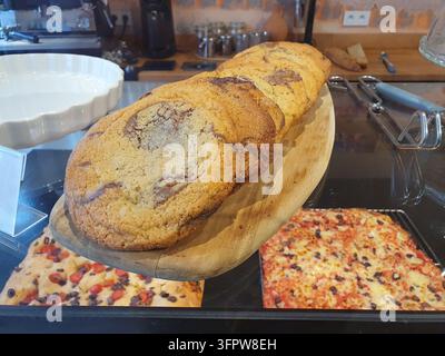 Große Schokoladenspiralkekse auf einem hölzernen Servierteller hinter einer gläsernen Bäckereitheke, darunter verschiedene Pizzen sichtbar, was eine warme  Stockfoto