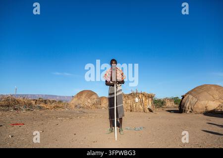 Älterer Afar-Mann steht mit einem Holzstock in der Nähe traditioneller Hütten und Berge, Afar-Region, Afambo, Äthiopien Stockfoto