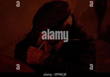 Atmosphärisches Straßenfoto bei schwachem Licht eines älteren Mannes mit flacher Kappe und dunkler Jacke, der unter warmem rotem Licht eine Zigarette anzündet. Stockfoto