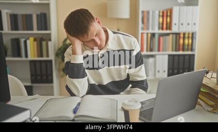 Junger Studenten, der seinen Kopf auf die Hand stützt, über offenen Büchern, Notizen und einem Laptop in einem Studienbereich eines Gebäudes; Studienmüdigkeit. Stockfoto