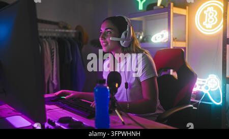 Frau setzt Headset auf, während sie nach Tastatur und Mikrofon im neonbeleuchteten Desktop-Studio greift, passt Headset in der Nähe des Monitors und des Gaming-Stuhls an; konzentriert Stockfoto