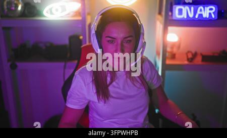 Frau mit Headset und weißem T-Shirt, sitzt in einem Gaming-Stuhl und blickt nach vorne unter einem Neon-On-Air-Schild im Studio; konzentrierte Entschlossenheit. Stockfoto