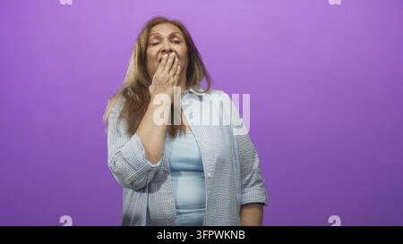 Frau in hellblauem Oberteil bedeckt den Mund mit der Hand in einem Studio-Porträt vor einem lila Hintergrund; überraschende Reflexion. Stockfoto