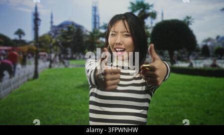 Junge Frau in gestreiftem Pullover macht mit beiden Händen ein OK-Zeichen und spitzt die Lippen, während sie auf grünem Rasen vor einem historischen Gebäude steht; verspielt Stockfoto