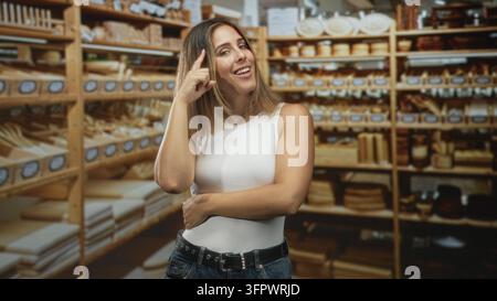 Frau zeigt mit dem Zeigefinger auf die Regale in einem Bastelstudio zwischen Holzschalen und Schneidebrettern, lächelt und bewegt selbstbewusst Stockfoto
