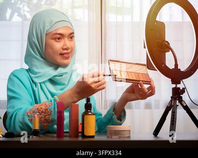 Glückliche junge asiatische muslimische Frau, die von zu Hause aus einen Beauty-Vlog streamt, Online-Content-Erstellerin, die ein Parfümduftmittel aufträgt Stockfoto
