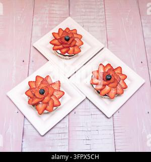 Drei blumenförmige Erdbeer-Dessert-Tartlets mit Schlagsahne und einem Blaubeerkern, auf quadratischen weißen Eco-Tellern über einem rosa abgenutzten Holztisch Stockfoto