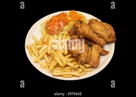 Knusprig gebratenes Brathähnchen mit gezackten Pommes, gerissenem Salat, Tomatenscheibe und geriebener Karottensalat auf einem runden weißen Teller vor bla Stockfoto