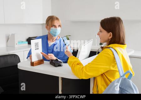 Kontaktloses Bezahlen und digitales Einchecken, Patient scannt QR-Code an der Empfangstheke einer medizinischen Klinik mit einer Krankenschwester, die eine schützende Gesichtsmaske trägt, um die Gesundheit zu schützen Stockfoto