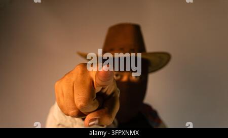 Kreatives Studio-Porträt eines jungen asiatischen Mannes mit dunkler Balaklava und braunem Cowboyhut. Die verkleidete Person zeigt mit dem Zeigefinger dire Stockfoto
