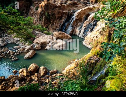 Ein kleiner Wasserfall fließt durch schroffe Felsen in ein klares türkisfarbenes Becken, umgeben von üppigem grünem Wald und sonnenbeschienenen Pflanzen. Stockfoto
