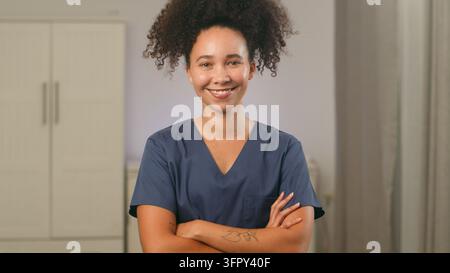 Gesundheitsarbeiterin lächelt mit verschränkten Armen, während sie im Innenbereich medizinische OP-Kleidung trägt Stockfoto