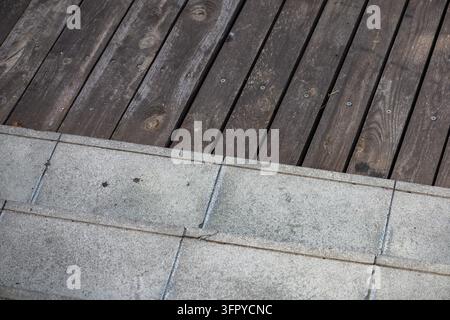 Graue Betonstufen treffen auf dunkelbraune, verwitterte Holzterrassenplanken und schaffen einen klaren, texturierten Oberflächenkontrast auf einer architektonischen Außenterrasse. Visi Stockfoto